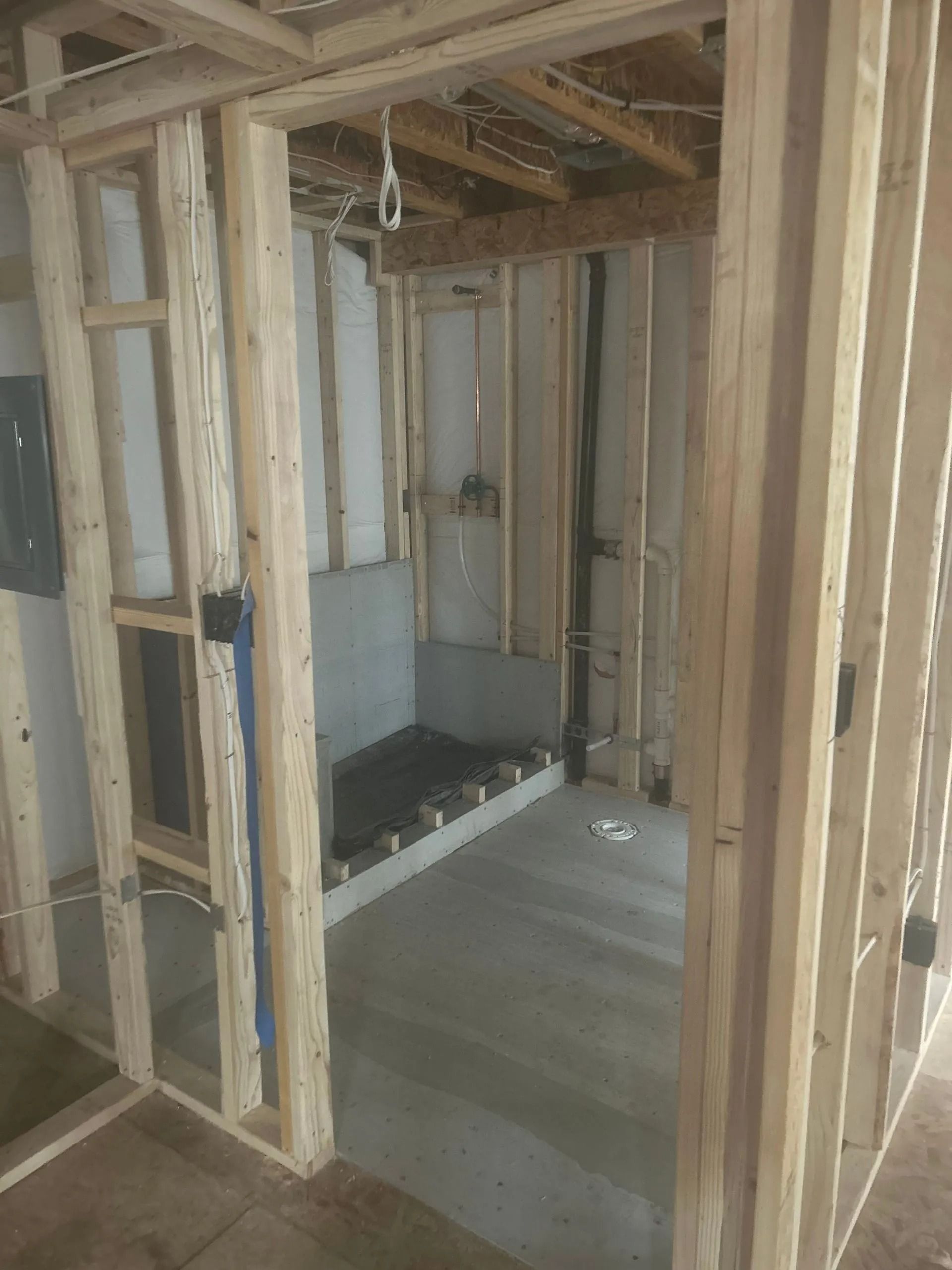 Framing of a bathroom under construction, showing studs, plumbing, and a concrete shower base.
