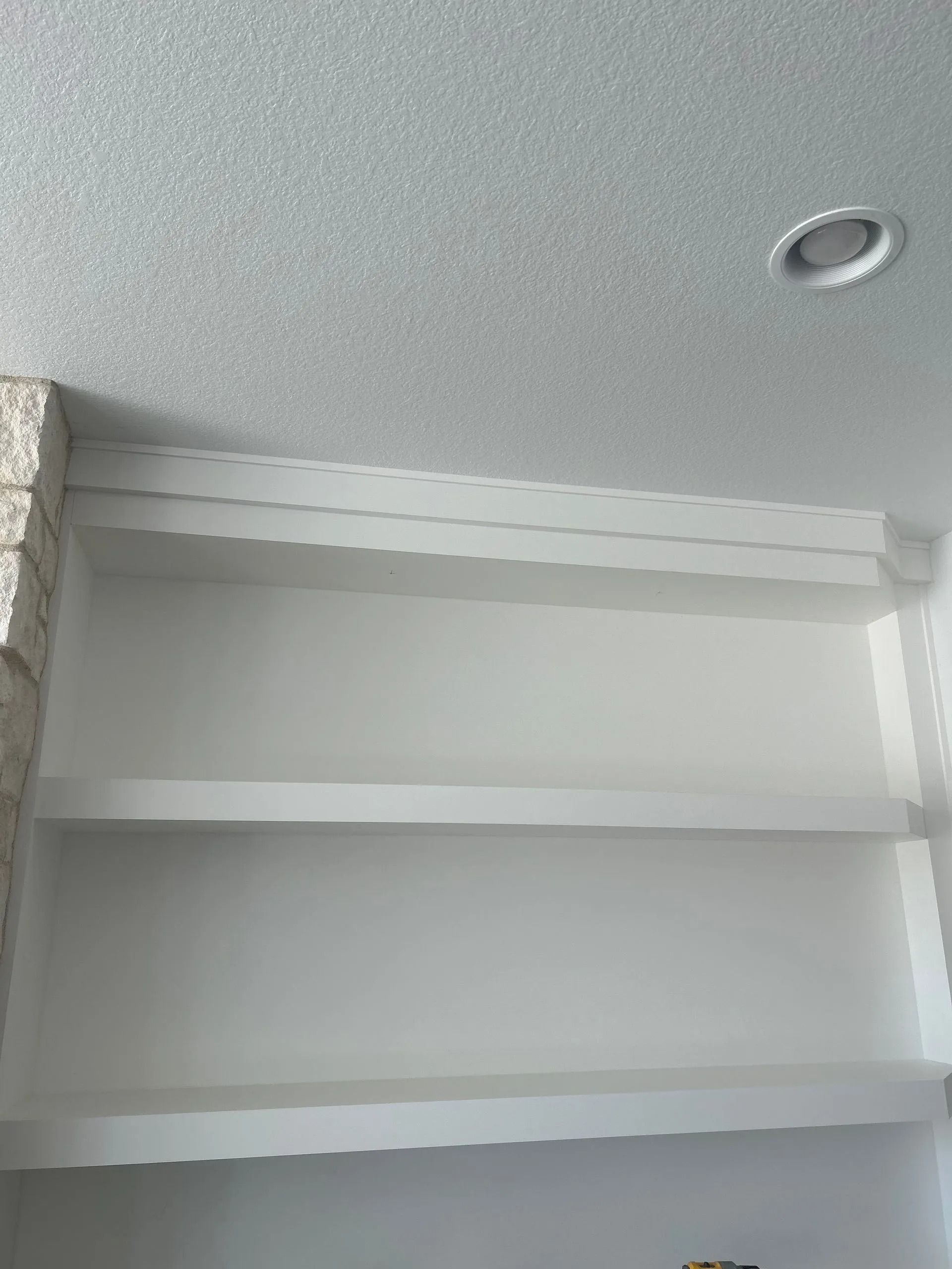 White built-in bookshelves with three shelves. Against a white ceiling. A recessed light is visible.