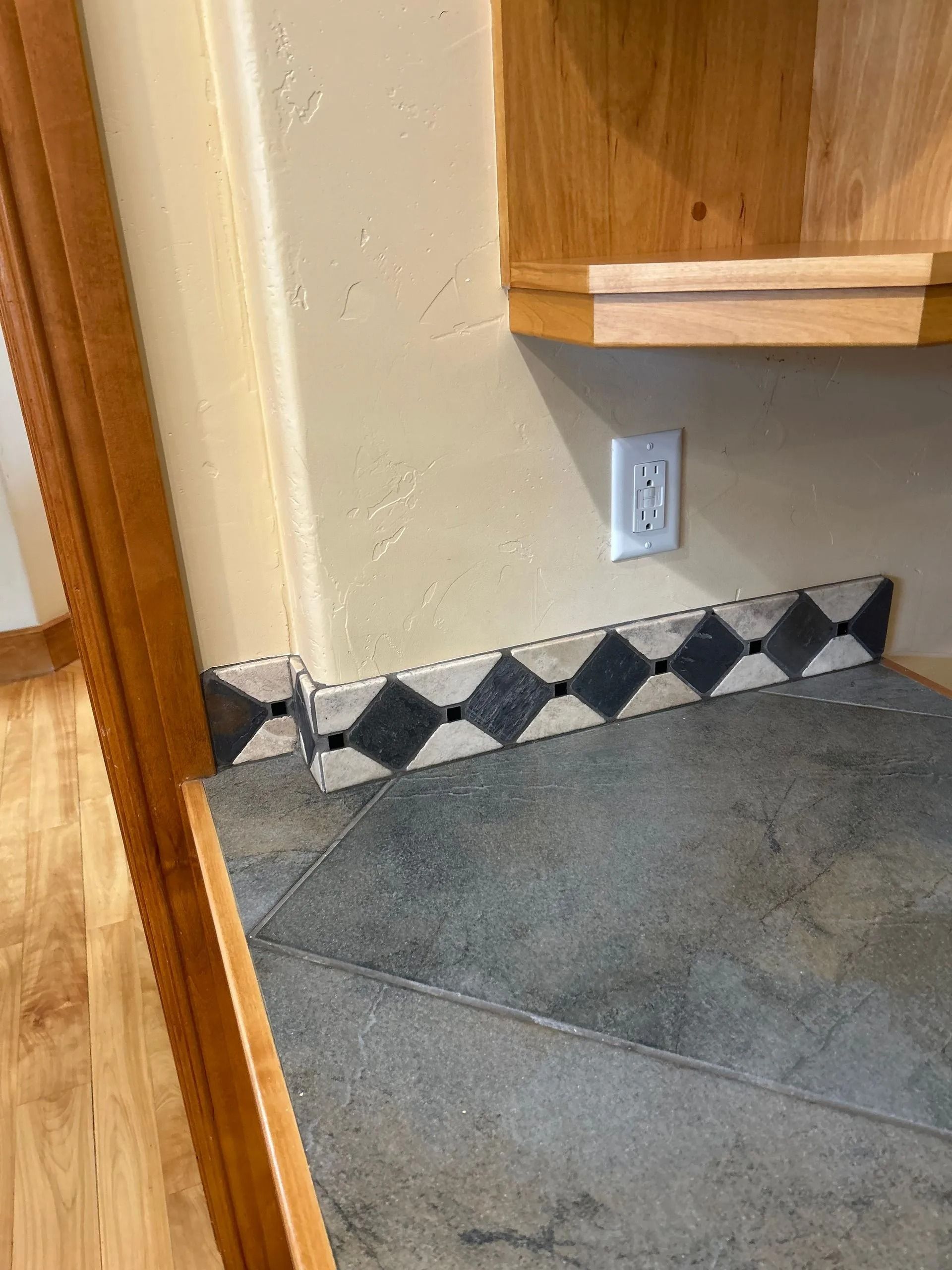 Corner of a kitchen counter with diamond-patterned tile backsplash. Wooden cabinets and a wall outlet are visible.