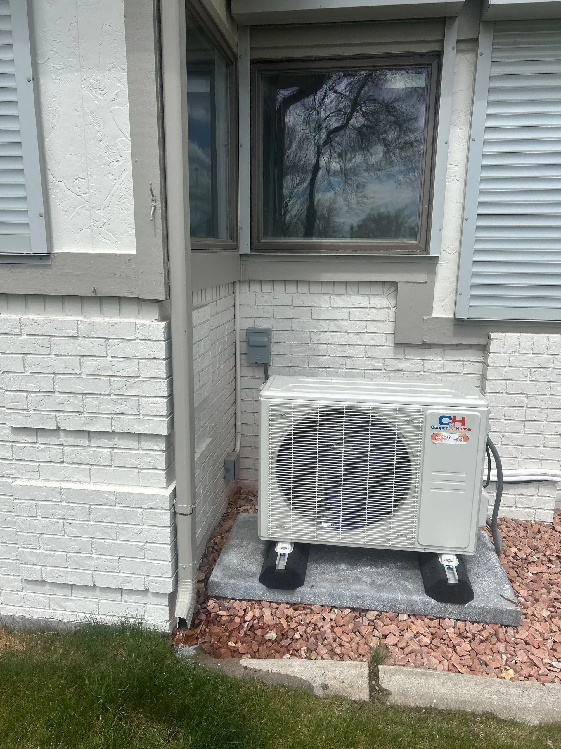 Air conditioning unit next to a house with white brick walls, under a window with shutters.