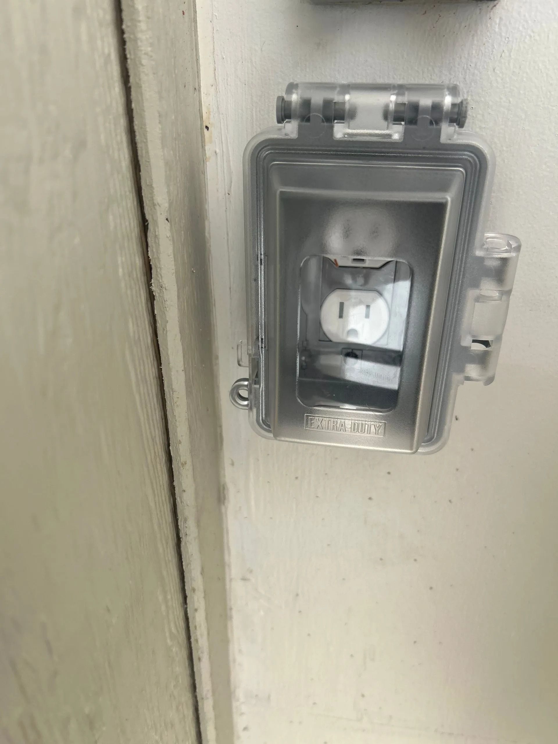 White outdoor electrical outlet with a clear protective cover on a white wall.