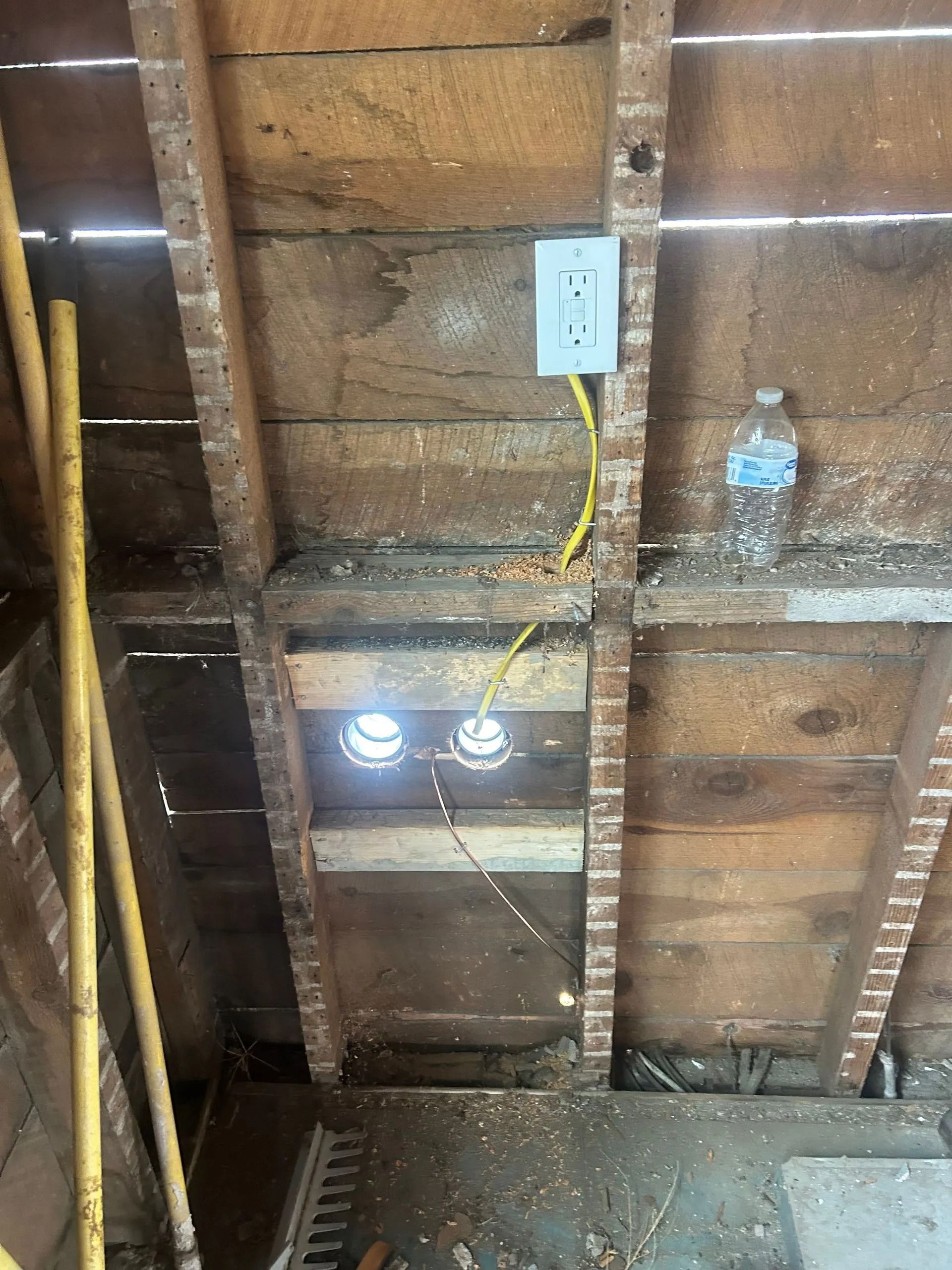 Overhead view of an unfinished attic space, showing wiring, electrical outlet, and two recessed light fixtures.