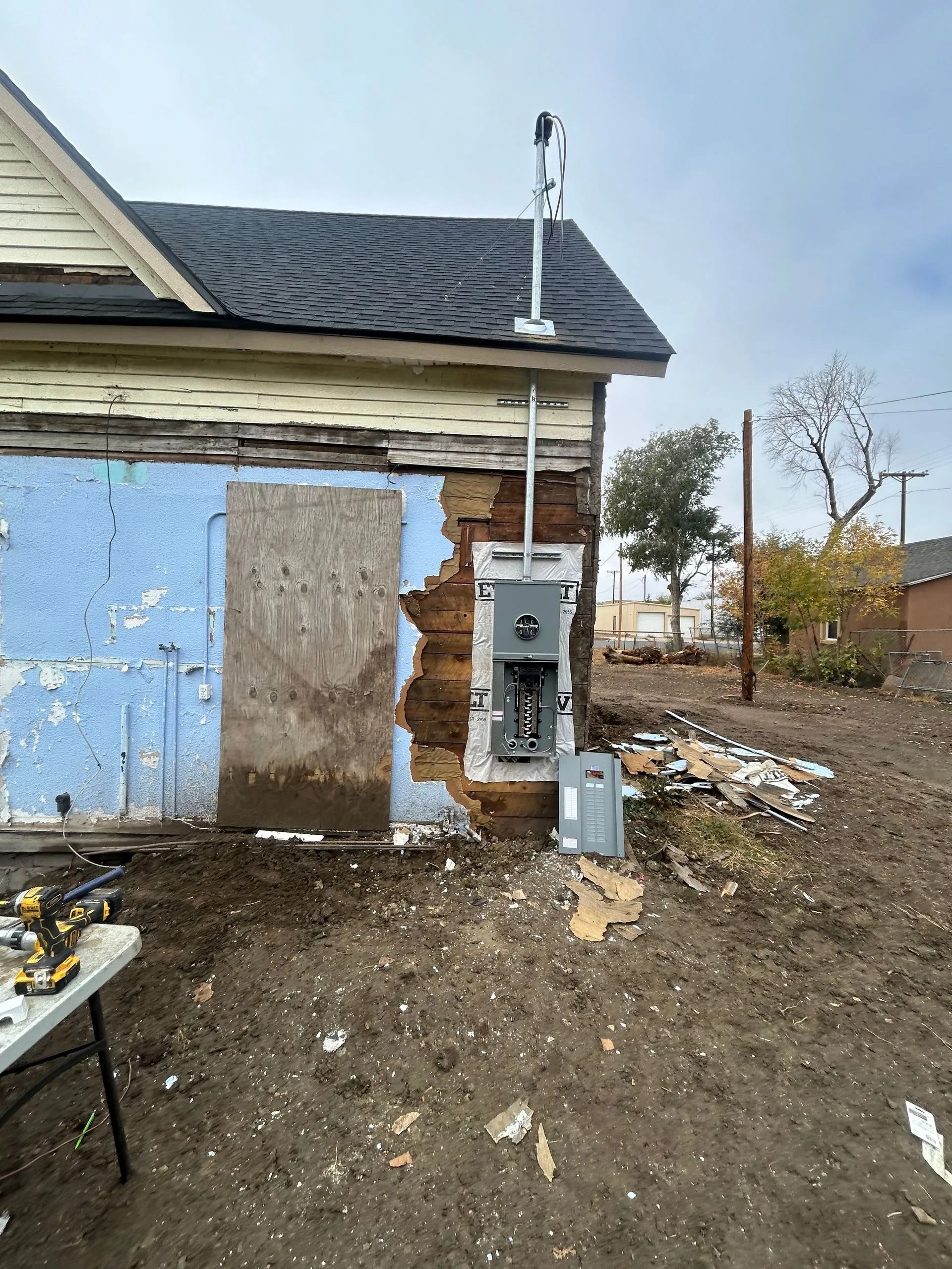 Exterior of a weathered house with exposed electrical panel and wiring. Construction debris on muddy ground.