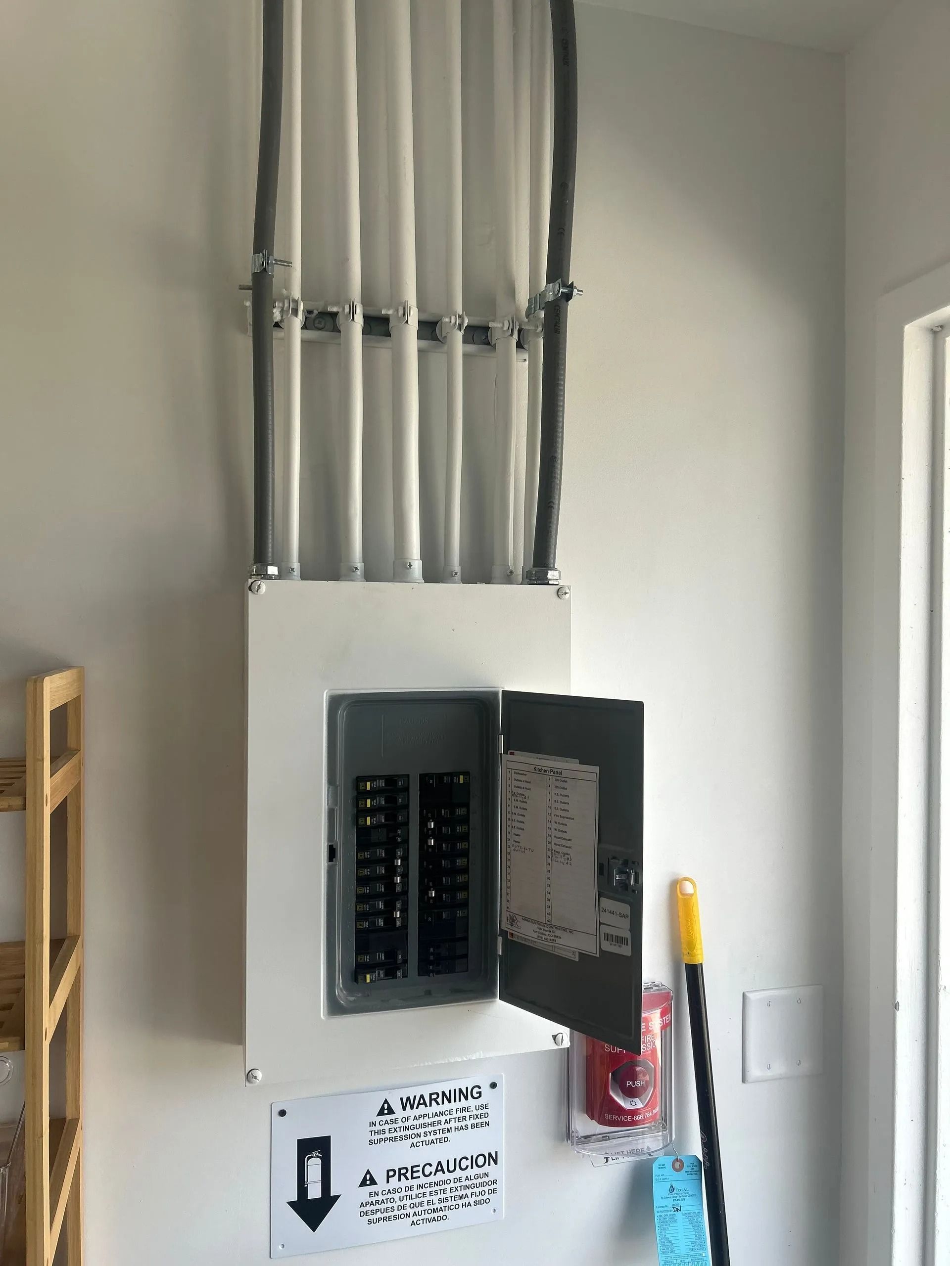 Electrical panel with open door, gray conduit pipes, and hazard warning sign on white wall.