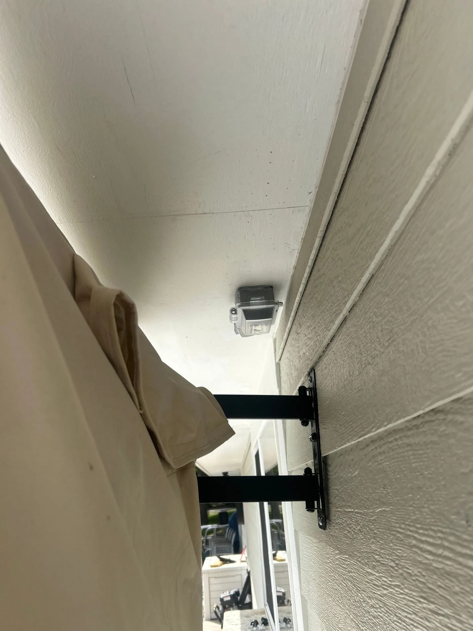 Black awning brackets mounted on a white wall, supporting a beige awning. A small device is mounted above.