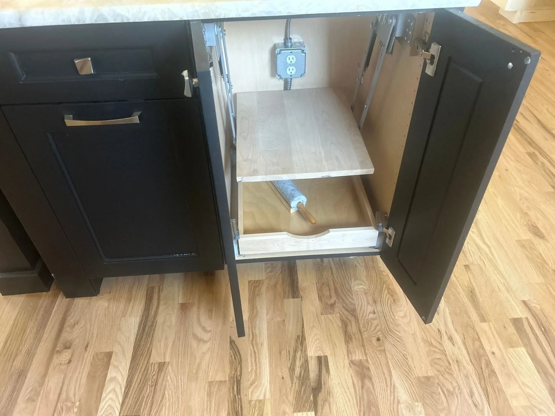 Black kitchen cabinet with pull-out shelf, open. Electrical outlet visible inside the cabinet.