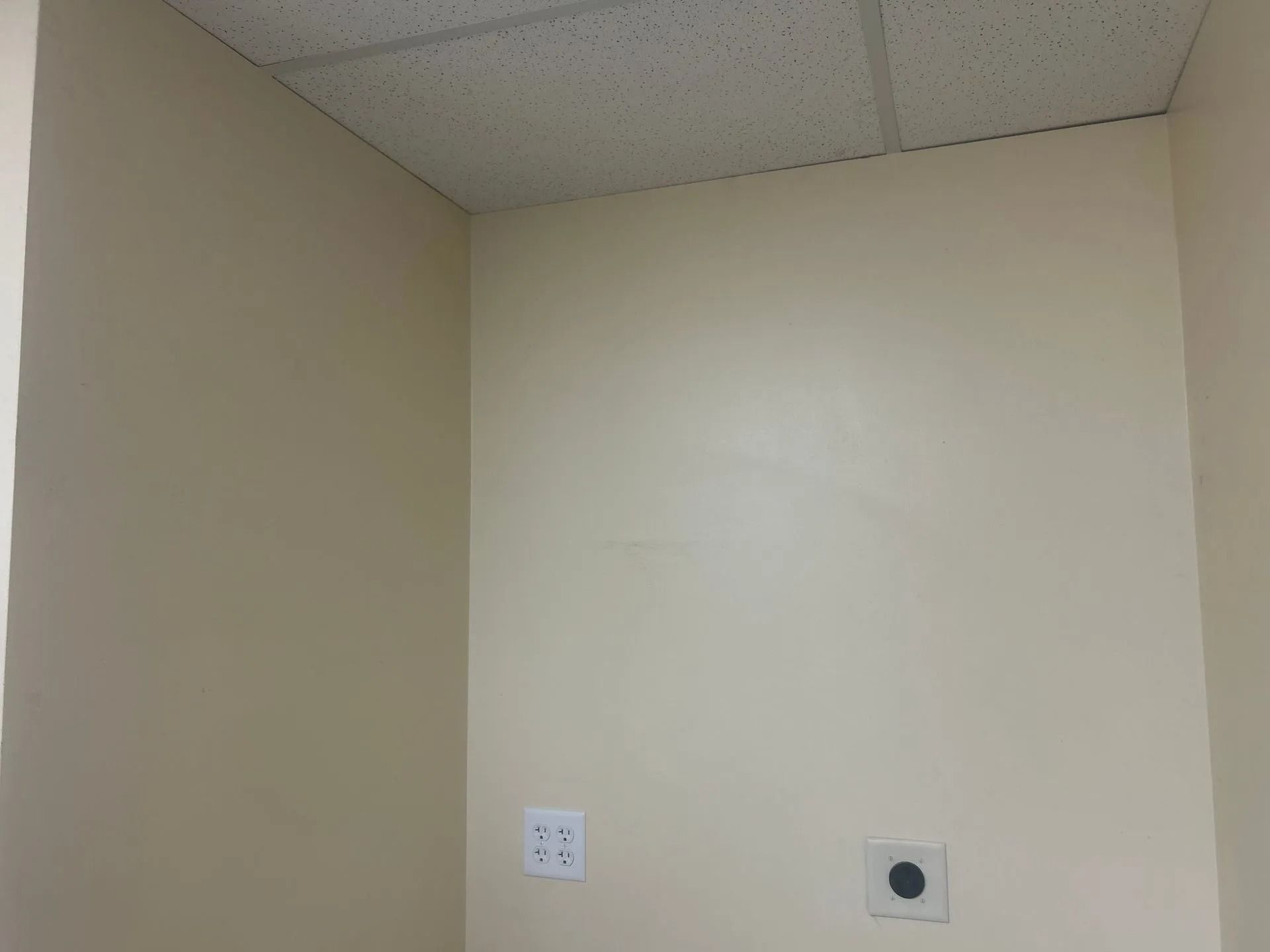 Corner of beige-painted room with textured ceiling, electrical outlet, and plumbing hookup on wall.