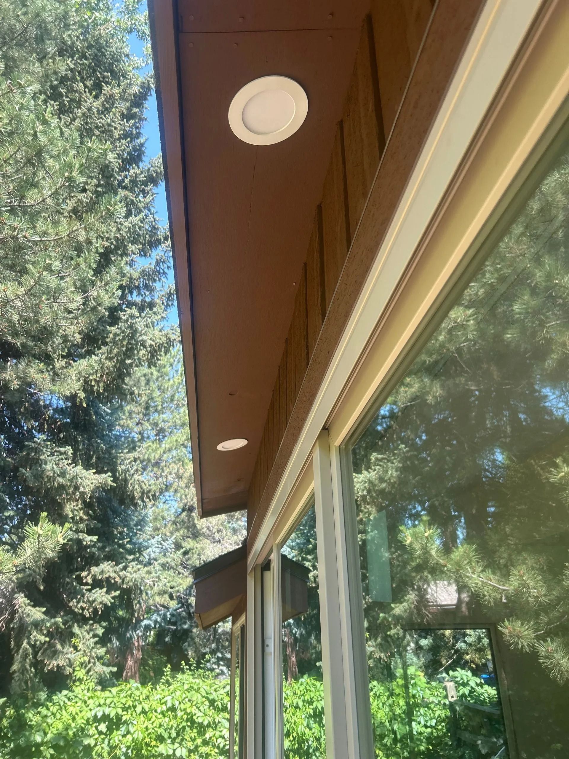 Brown eaves with white recessed lights above a window reflecting green trees.