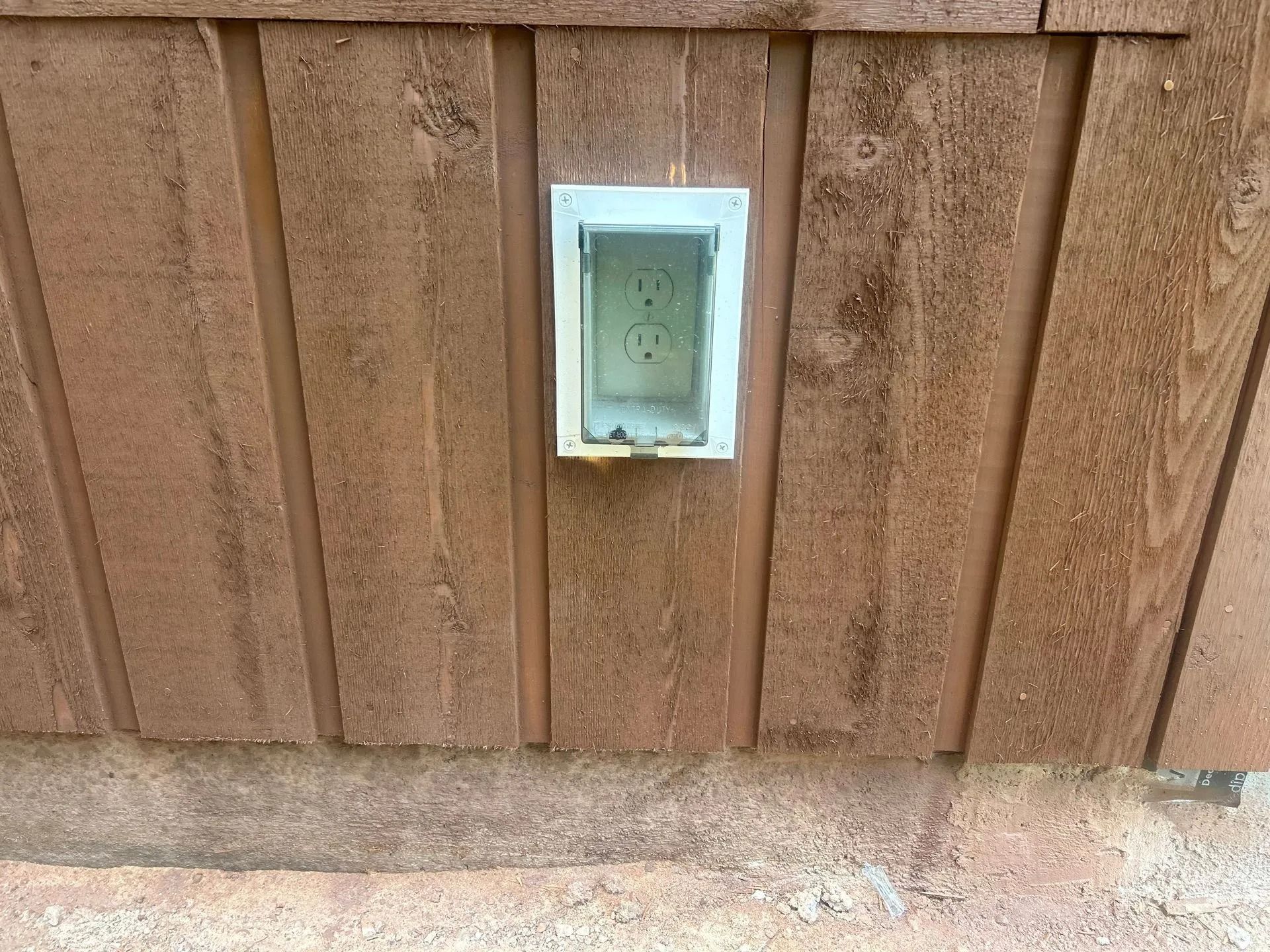 Outdoor electrical outlet in a white weatherproof enclosure on a brown wooden wall.