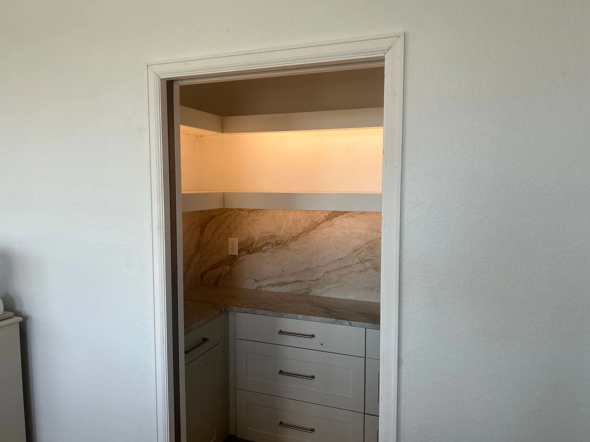 A pantry with white cabinetry, marble backsplash, and shelves lit by warm light, framed by a doorway.