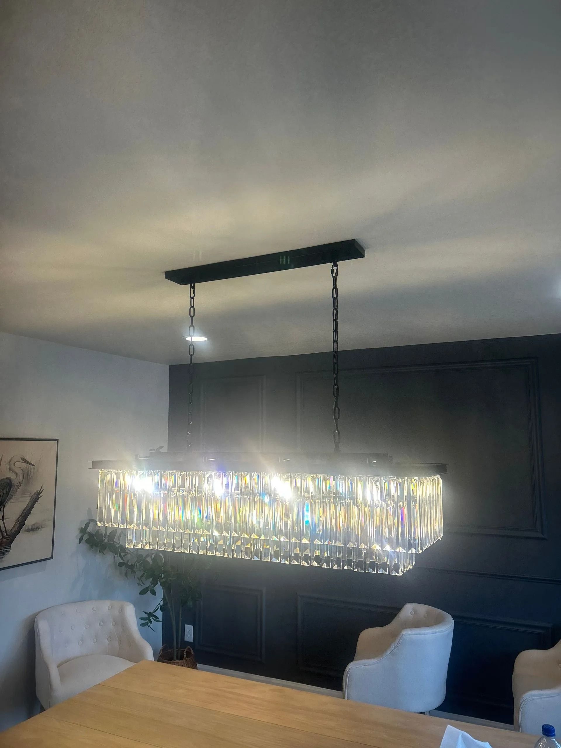 Crystal chandelier over a dining table with white chairs and a dark wall.