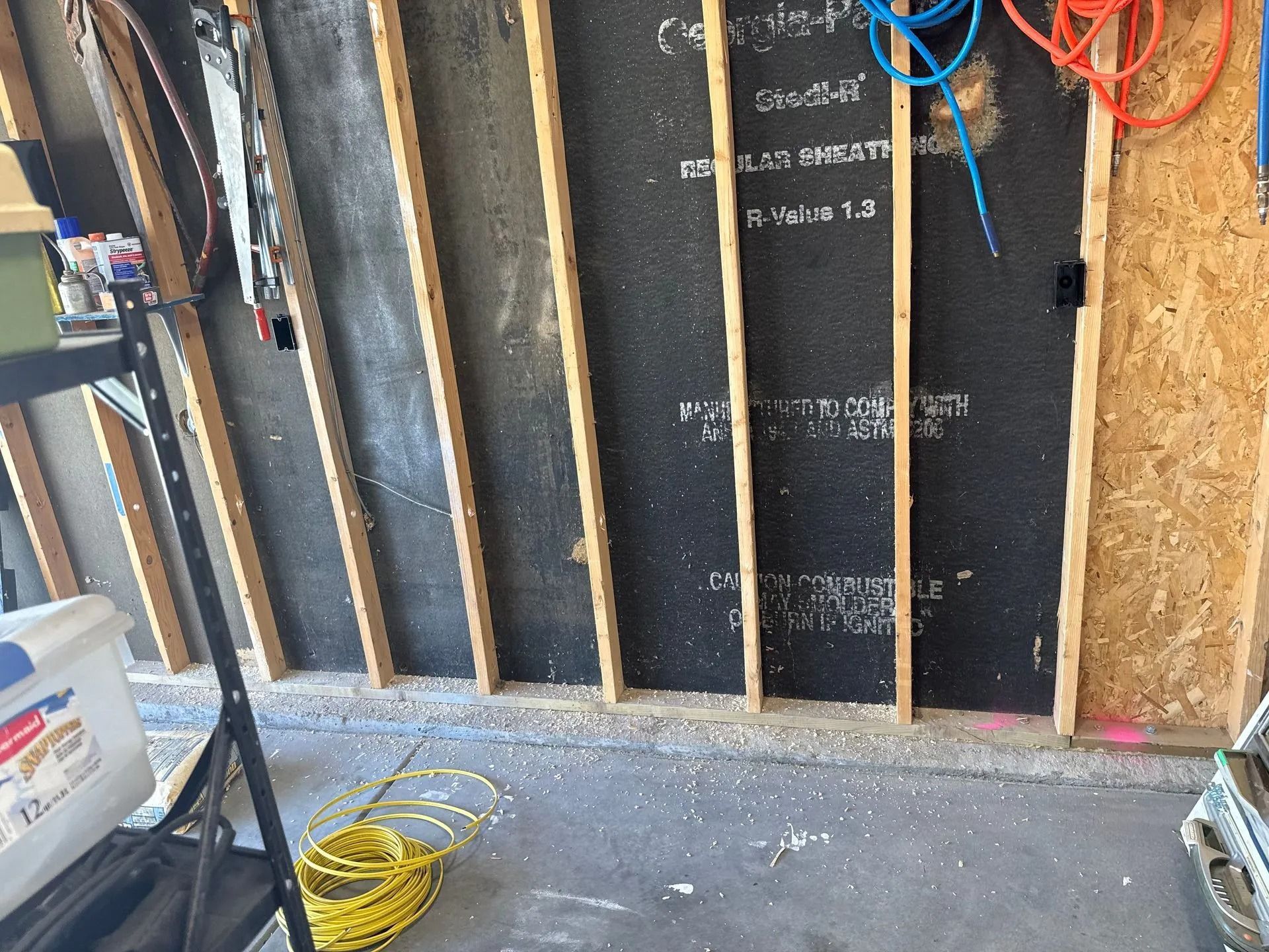 Garage wall with exposed studs and black sheathing. Various tools and equipment are visible.