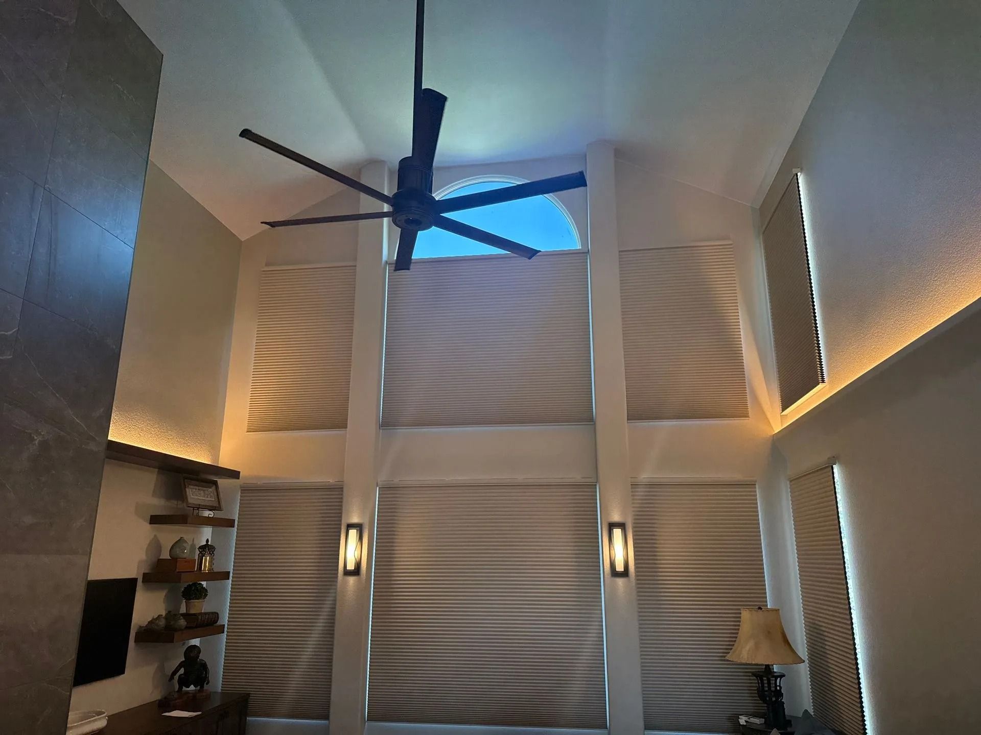 High-ceilinged living room with a large fan, tall windows with shades, and recessed lighting.