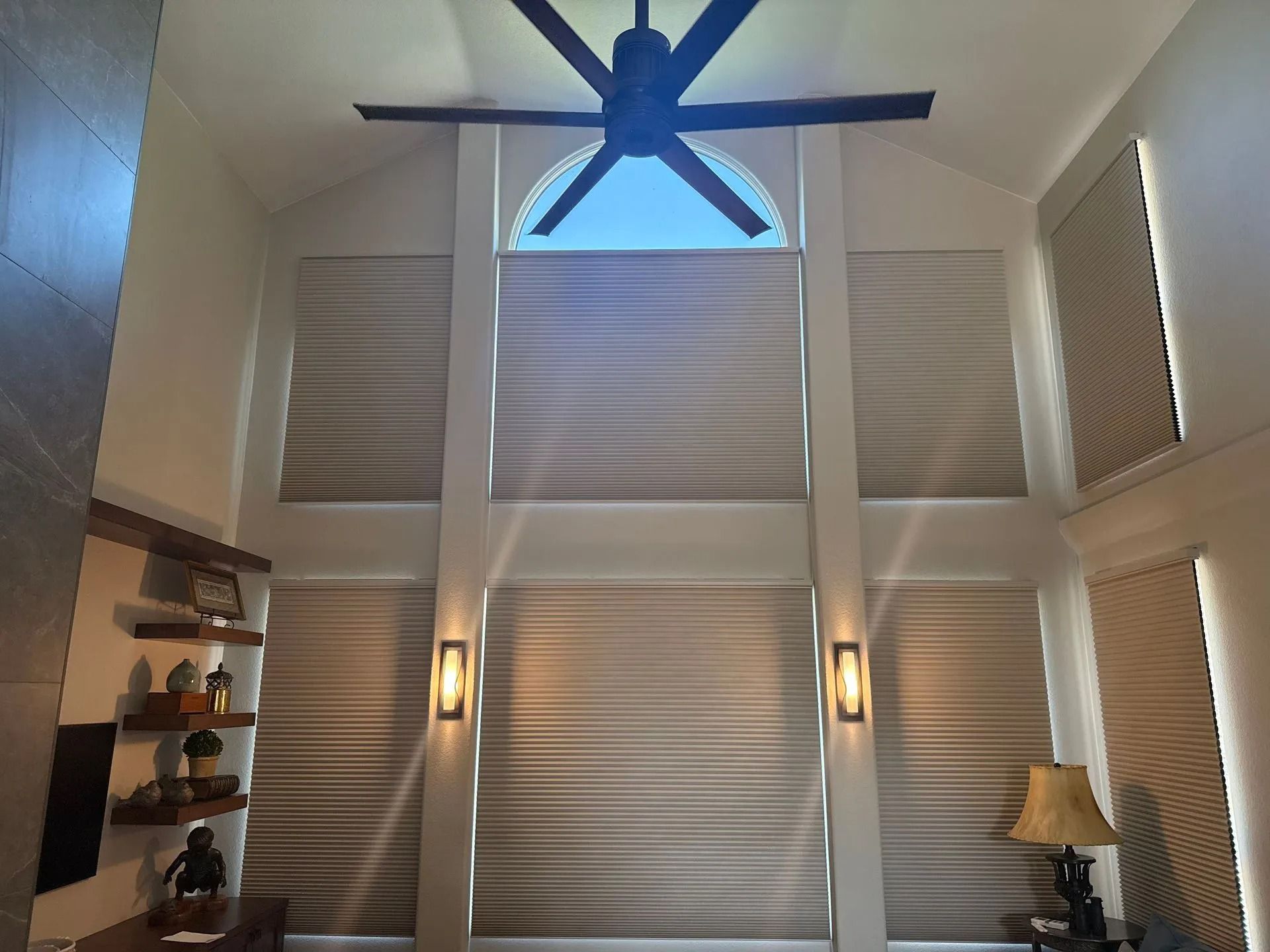 High-ceilinged room with window shades, ceiling fan, and wall sconces. Sunlight streams in.