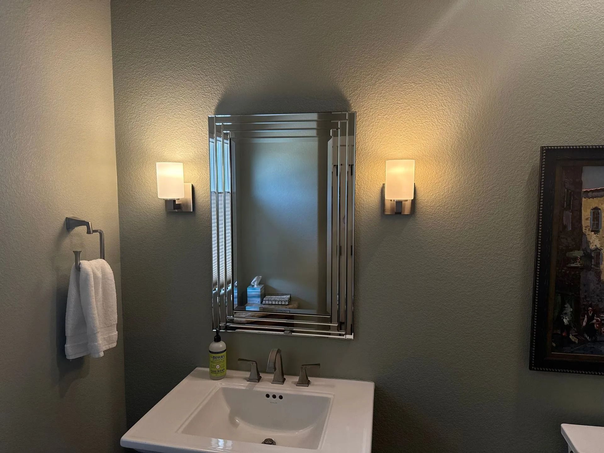 Bathroom with sink, mirror, sconces, and artwork on green textured walls.