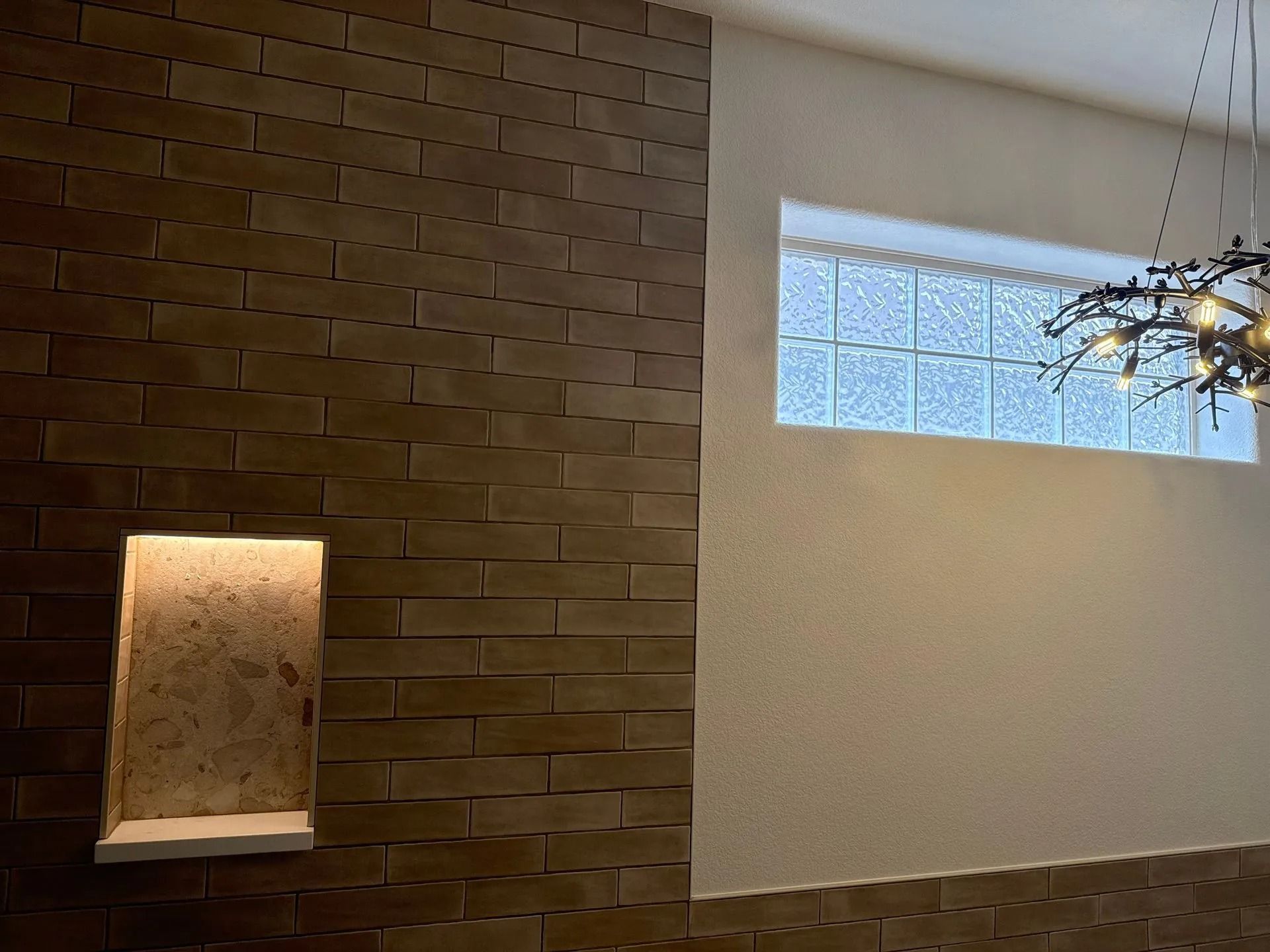 Brick wall with illuminated alcove next to a textured wall with a glass block window and a decorative light fixture.