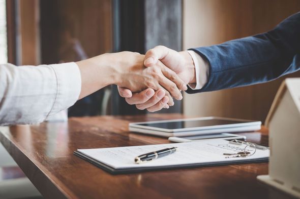 Two people shaking hands over a wooden table, with documents and a pen.