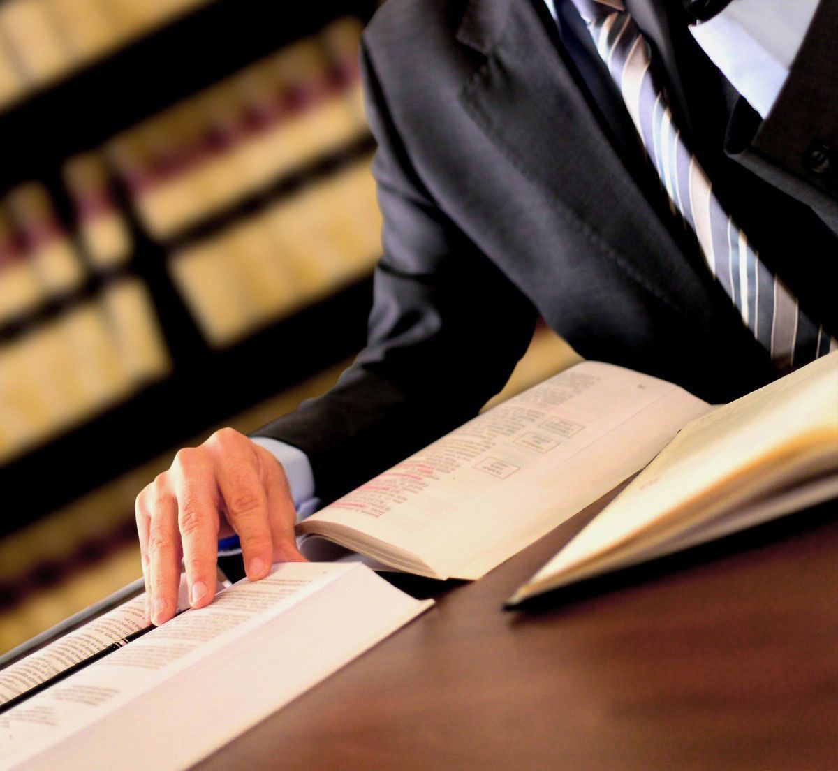 Man in suit, hand holding pen, studying a book at a desk with a bookshelf in the background.