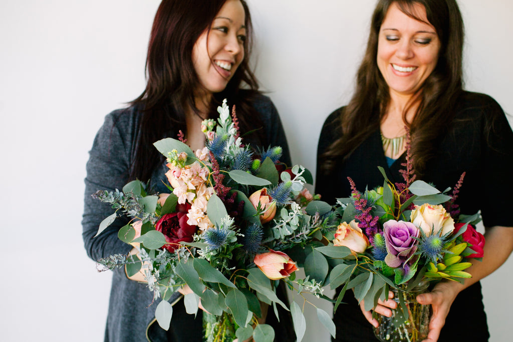 Two women smiling, holding colorful bouquets of flowers against a white background.