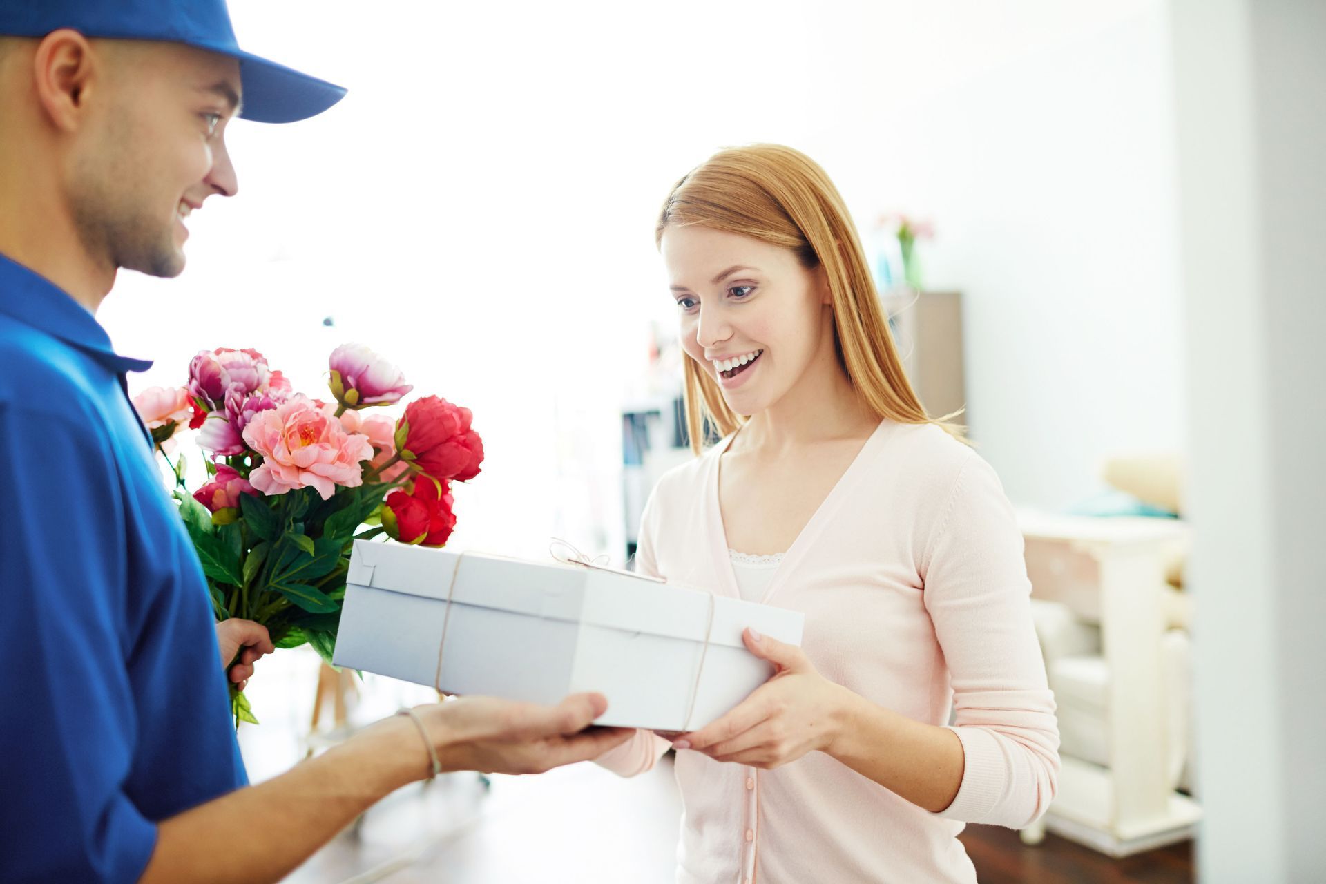 Delivery person hands flowers and a gift box to a smiling woman indoors.