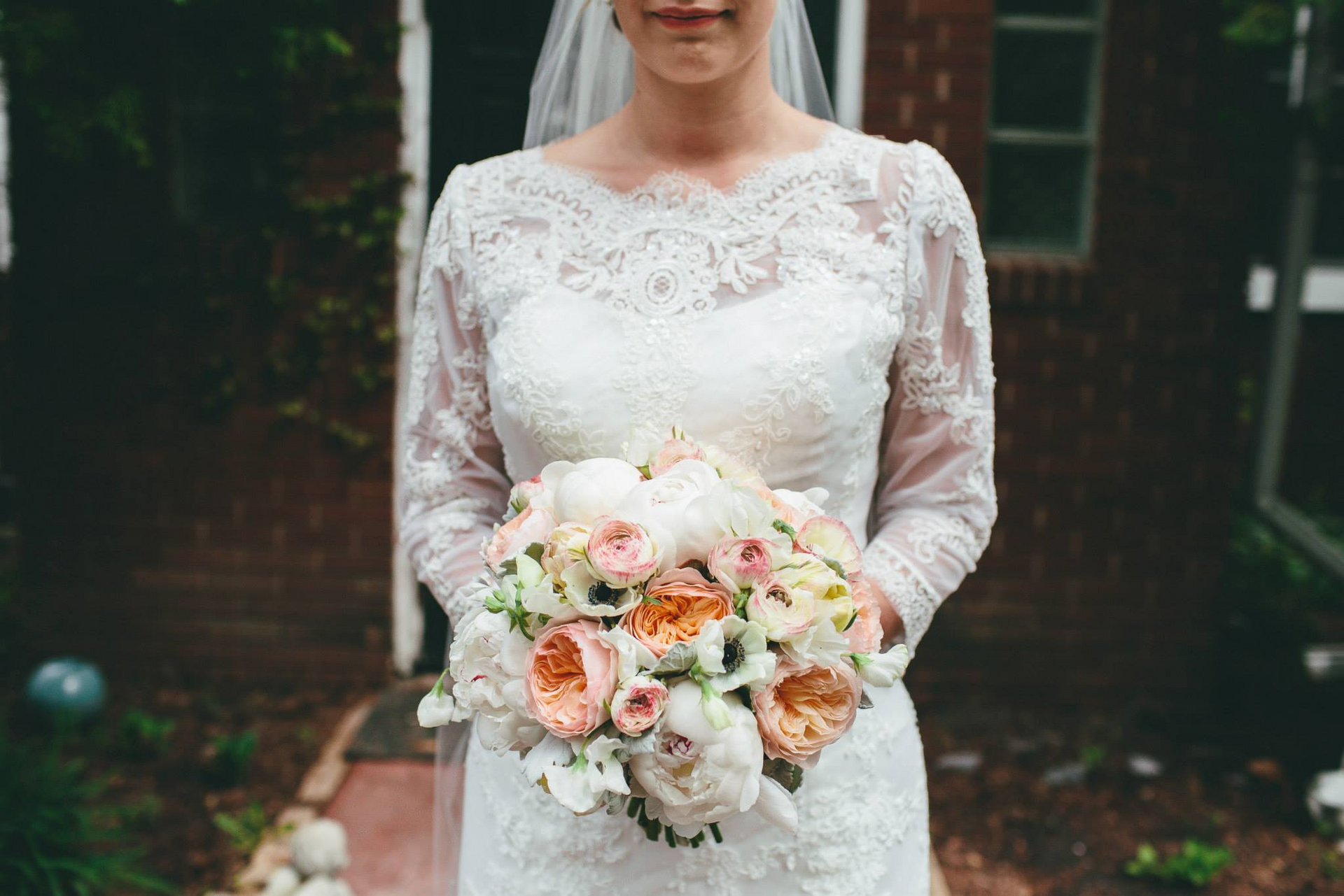Bride in a lace gown holds a bouquet of pink and white flowers outdoors.