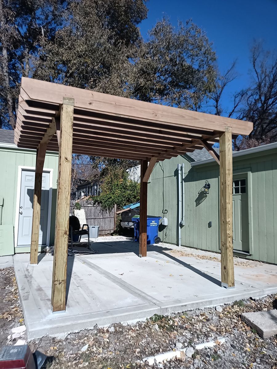 A wooden pergola is sitting on top of a concrete patio in front of a green building.