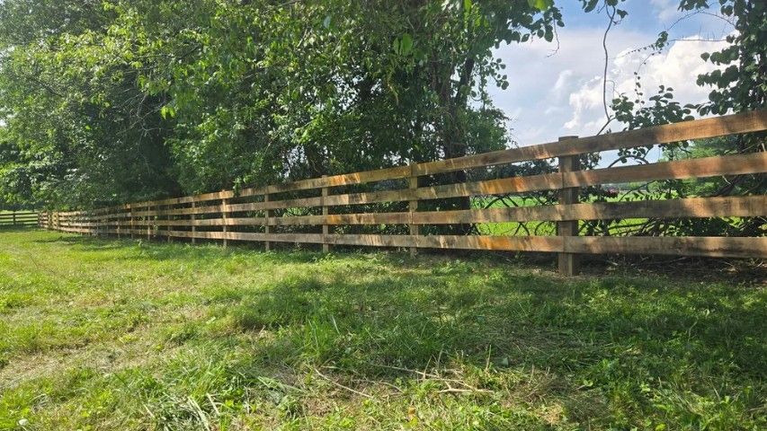 Wooden fence in a grassy field, shaded by trees under a partly cloudy sky.