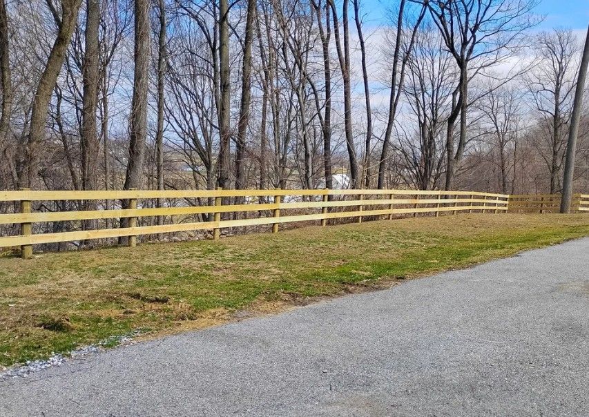 A long, light brown wooden fence stretches along a grassy area, bordering a gravel driveway and a wooded area.