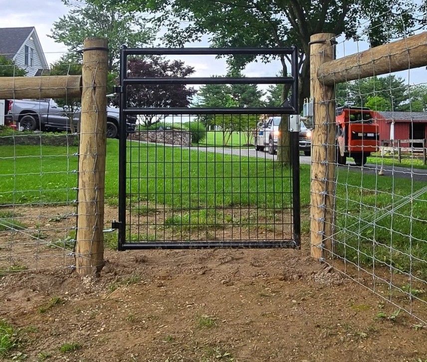 Black metal gate in a wire fence, flanked by wooden posts. Green grass and a house are visible in the background.