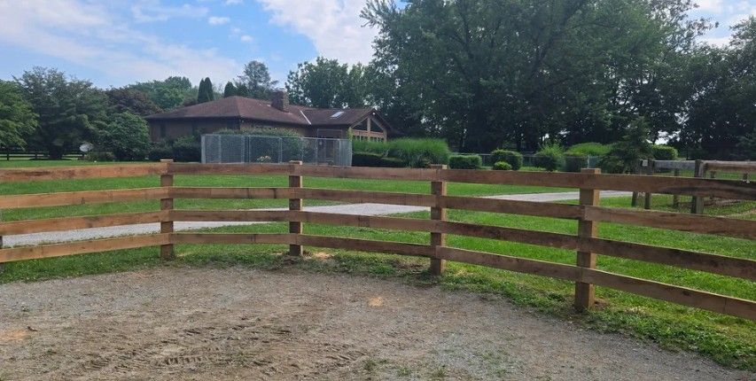 Wooden fence surrounding a gravel area, with a house and trees in the background on a sunny day.