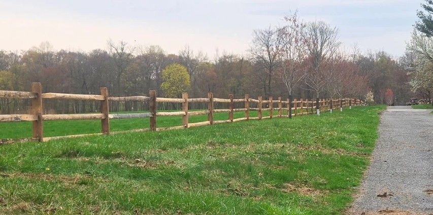 A split-rail fence borders a grassy field and a gravel path. Trees with autumn-toned leaves stand in the background under a cloudy sky.