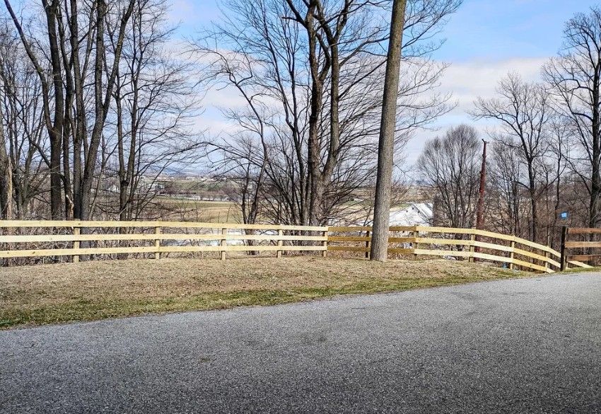 A wooden fence borders a grassy area next to a paved road, with leafless trees and a building in the distance under a blue sky.