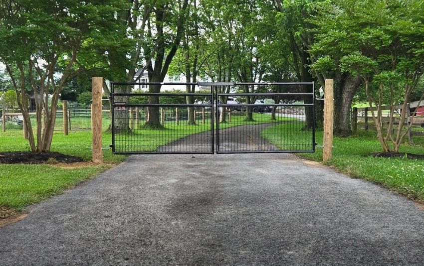 Driveway leading to a black metal gate flanked by wooden posts. Trees and a grassy area are visible beyond the gate.