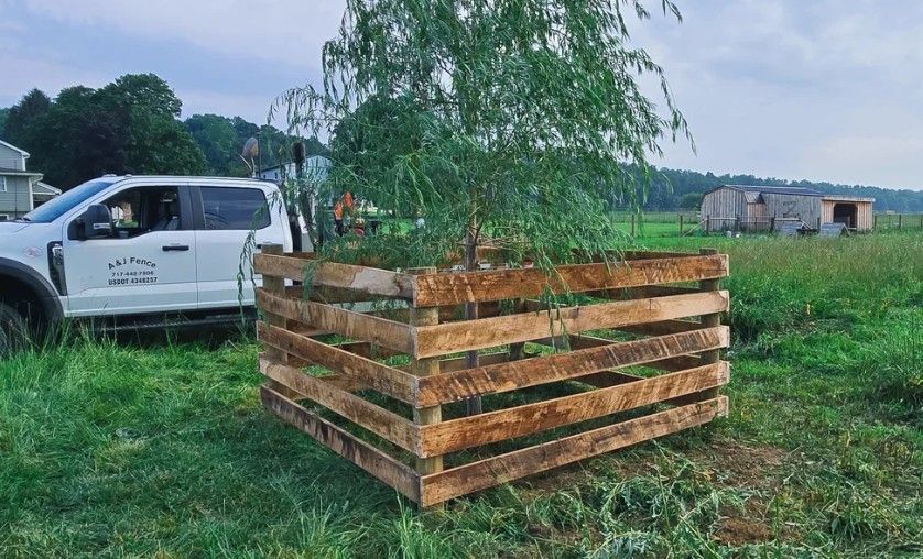 A young tree is protected by a wooden pallet fence in a grassy field, with a white truck parked nearby.