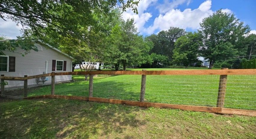 A wooden fence surrounds a grassy yard next to a white house with brown shutters, under a partly cloudy blue sky.