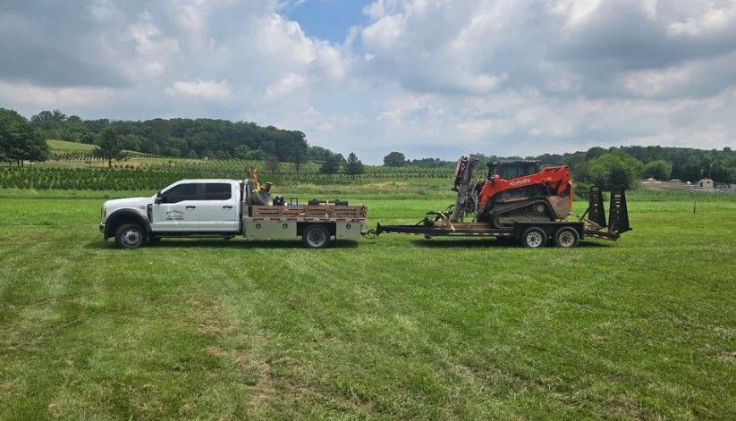 A white truck towing a trailer with an orange skid steer on a grassy field under a cloudy sky.