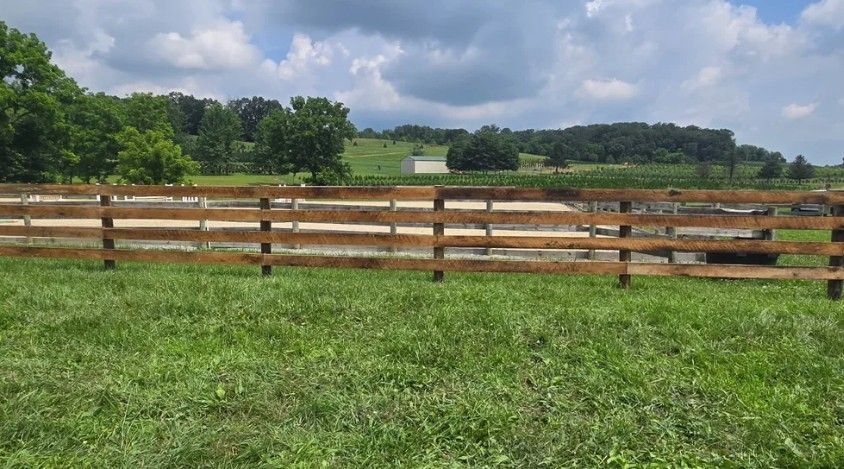 A wooden fence in a grassy field, with trees and a cloudy sky in the background.