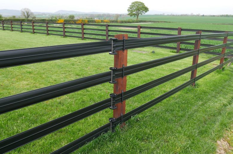 A black, multi-rail fence in a green field, with brown posts. A tree and hills are in the background.