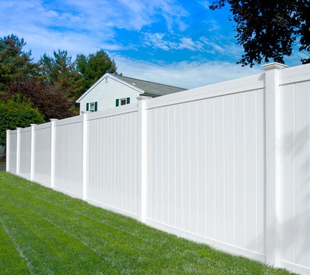 White vinyl fence surrounding a grassy lawn, with a house and trees in the background under a blue sky.