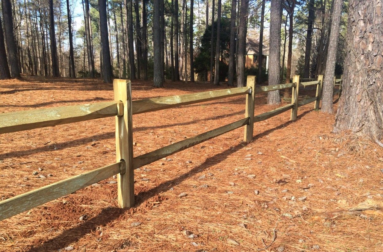 Wooden split-rail fence in a wooded area with a brown ground covered in pine needles.