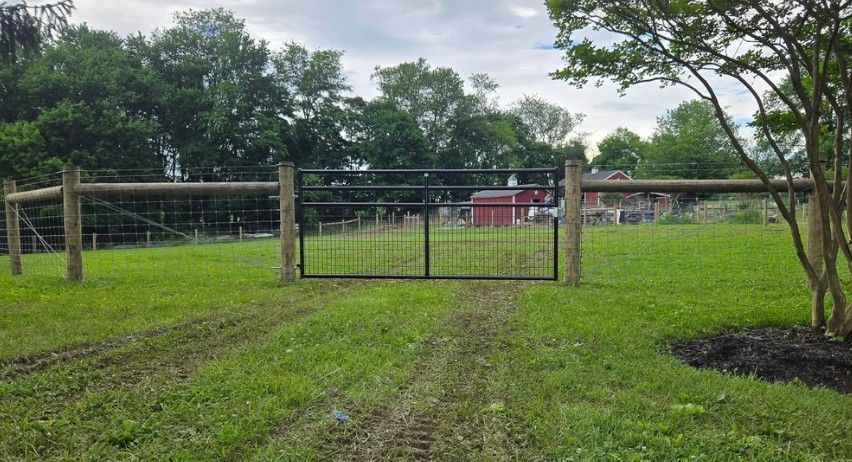 A metal gate in a green pasture leads to a red building, surrounded by a wooden fence and trees under an overcast sky.