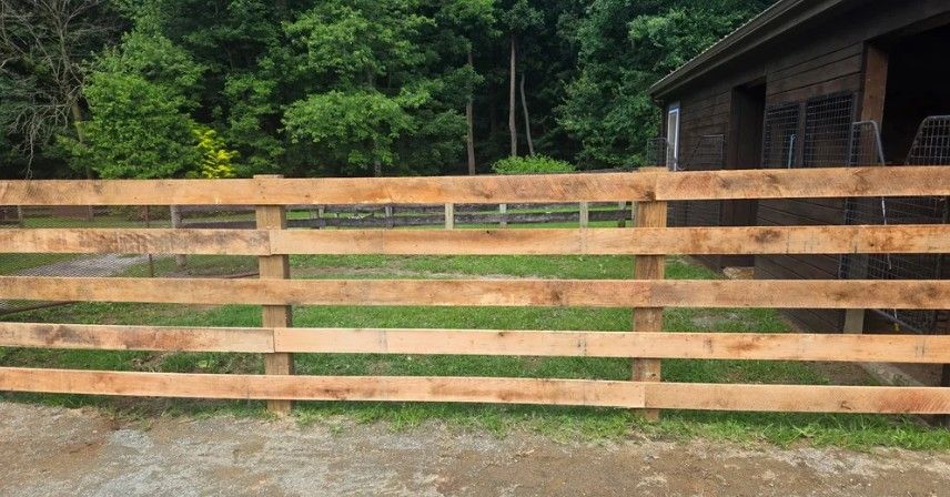 Wooden fence in front of a grassy area and trees, with a building visible in the background.