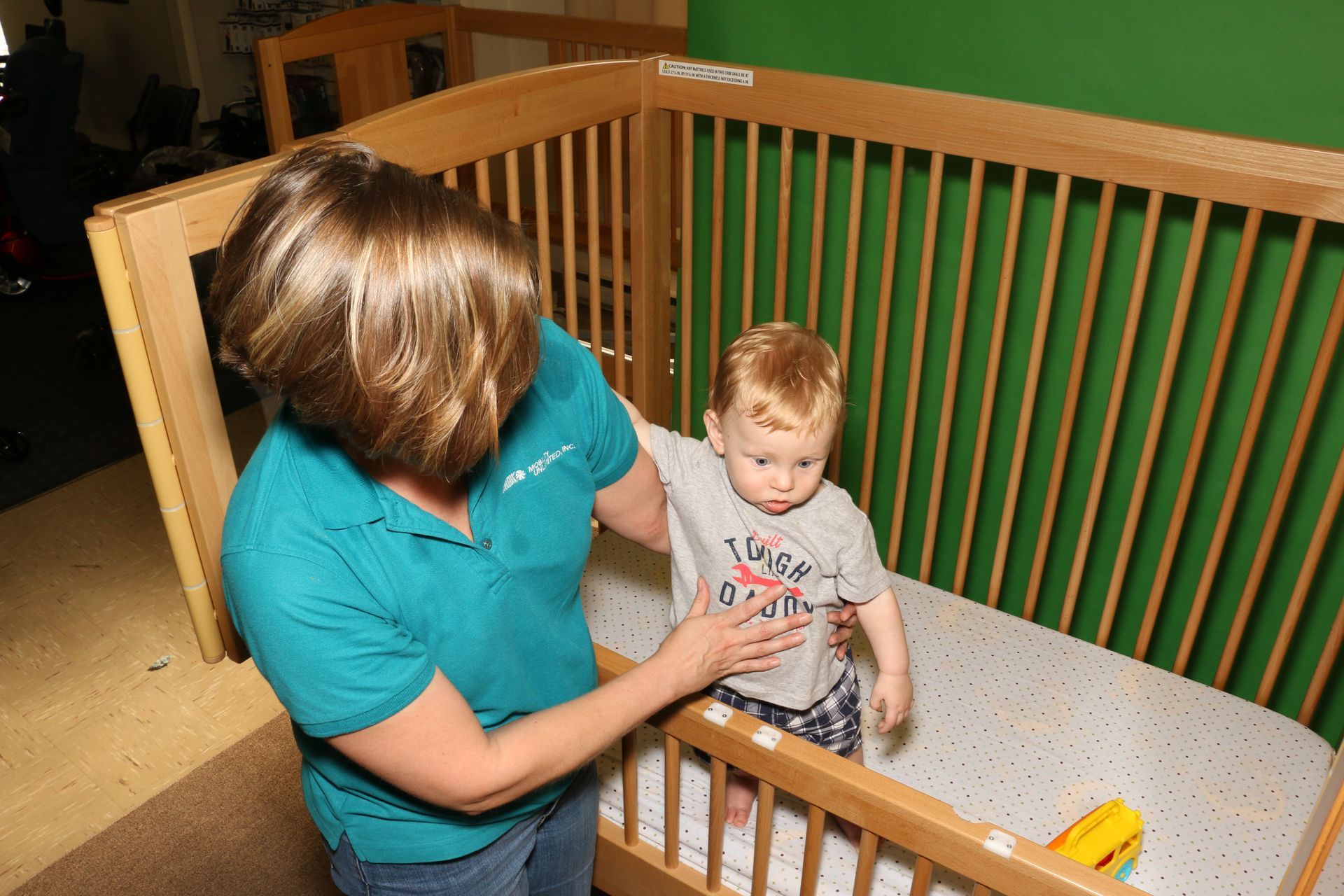 A woman in a teal shirt is helping a toddler out of a wooden crib in a room with green walls.