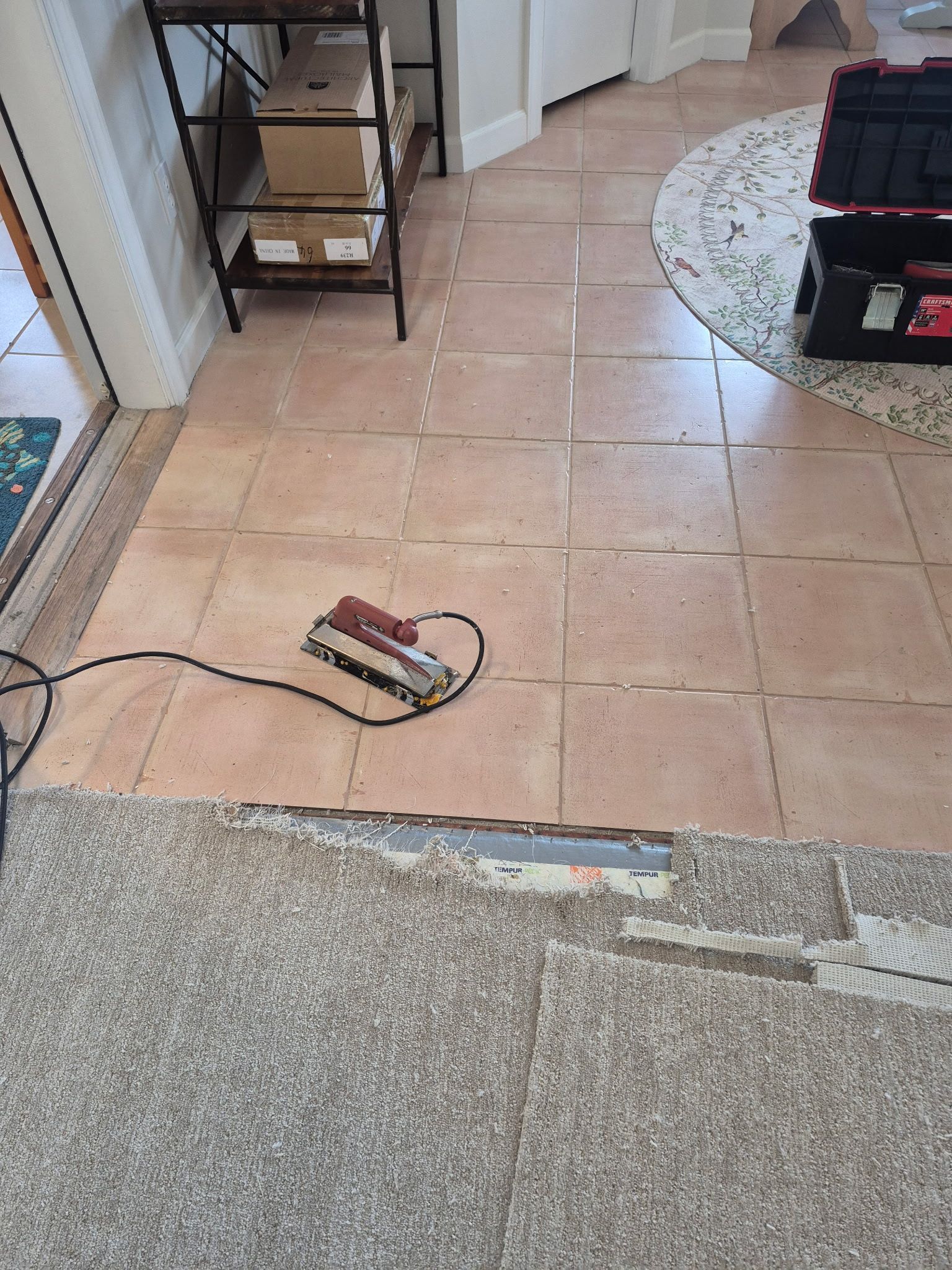 Hallway with loose floor tiles and a small battery pack on the tiled floor beside carpet edging