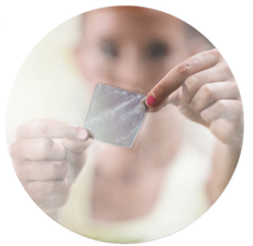 A person holds a small, clear, textured adhesive patch between their fingers against a blurred background.