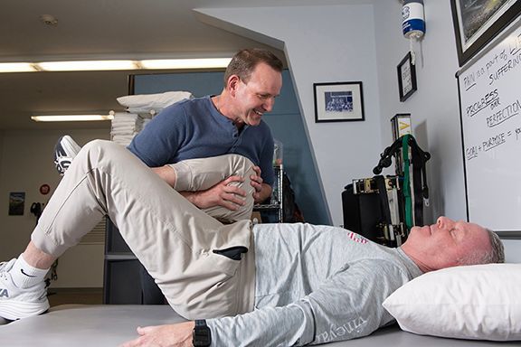 A physical therapist in a blue shirt stretches a client’s leg while the client lies on a table in a clinic.