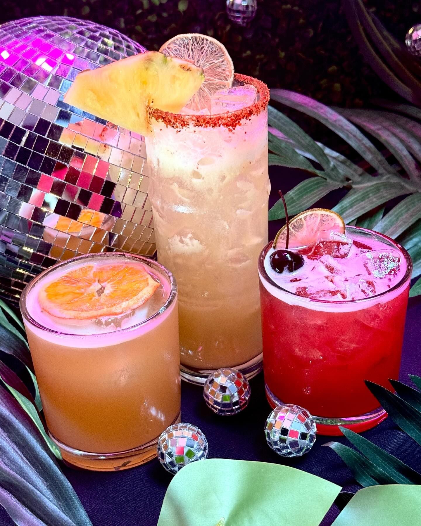 Three colorful cocktails with garnishes, next to a disco ball and tropical foliage.