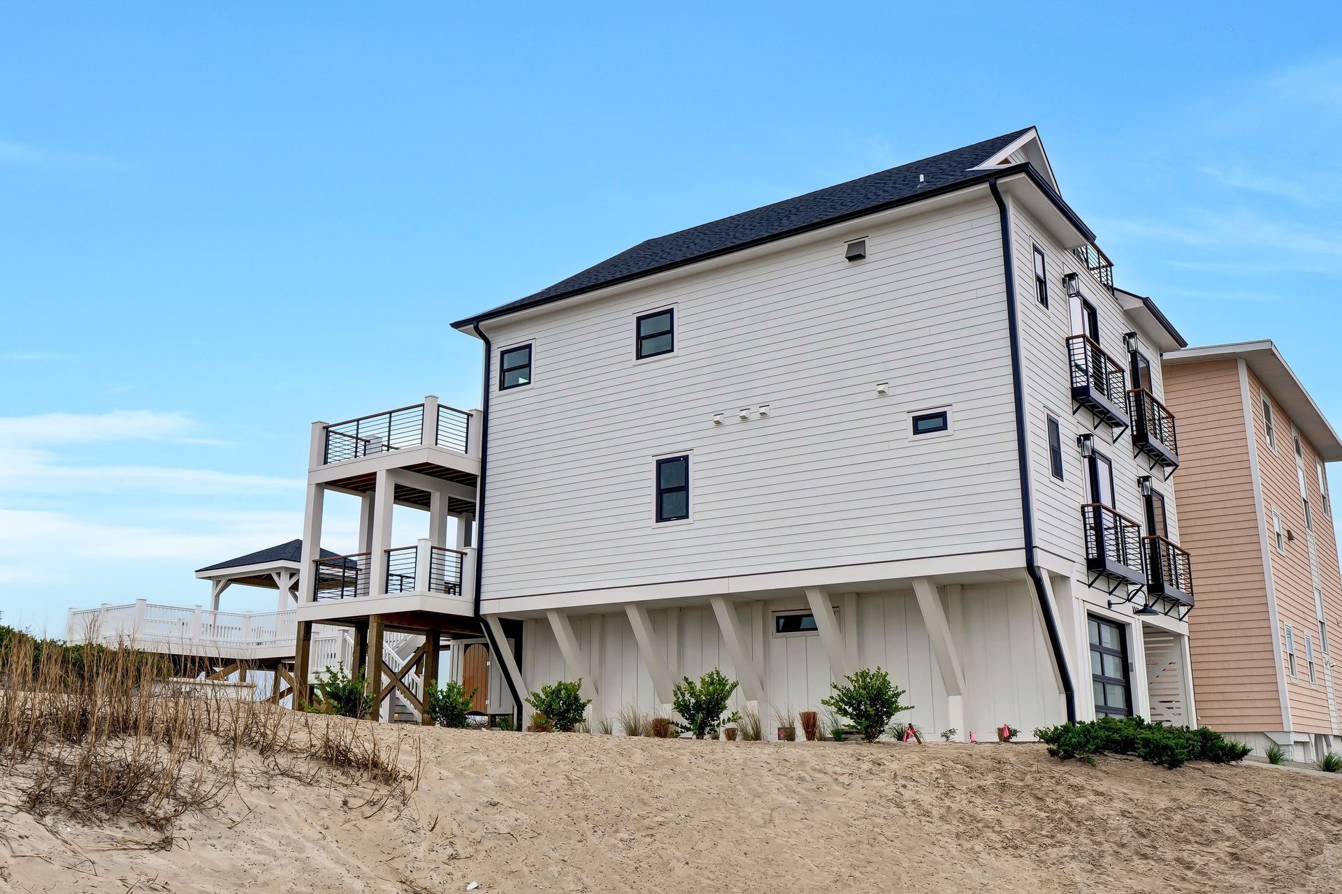 White beach house on a sand dune, blue sky. Balconies, dark roof, some shrubbery.
