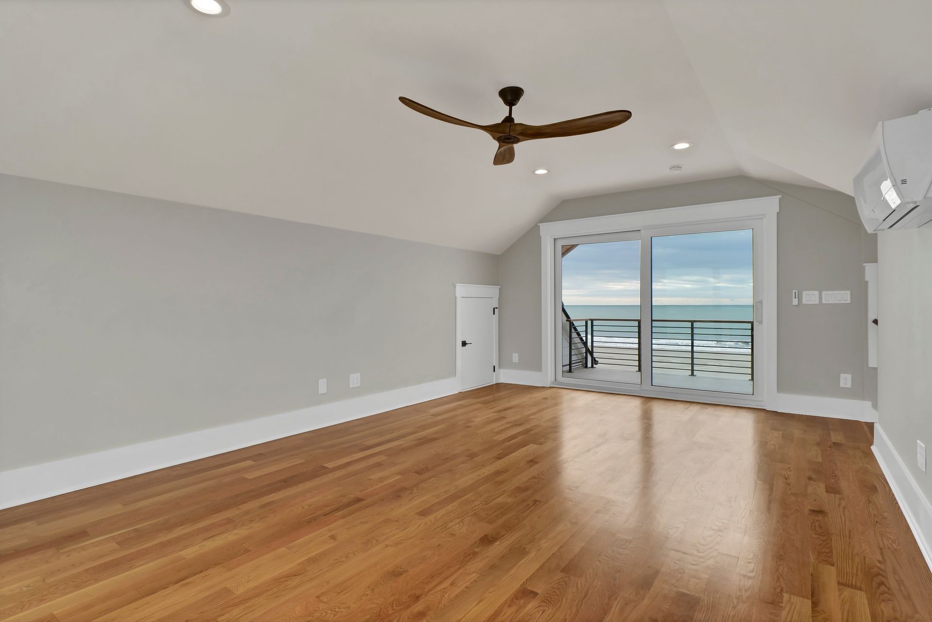 Empty room with hardwood floors, light gray walls, large window with ocean view, and ceiling fan.