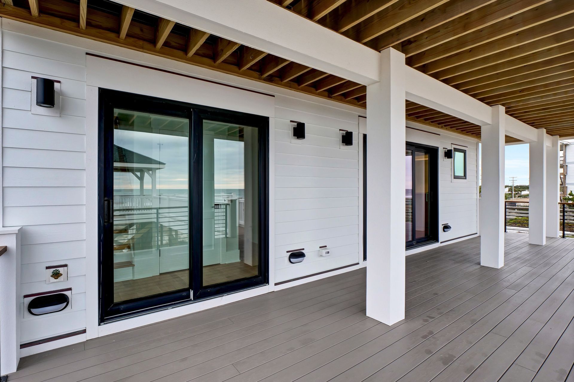 White beach house deck with black-framed glass doors and gray deck boards, under a wooden ceiling.